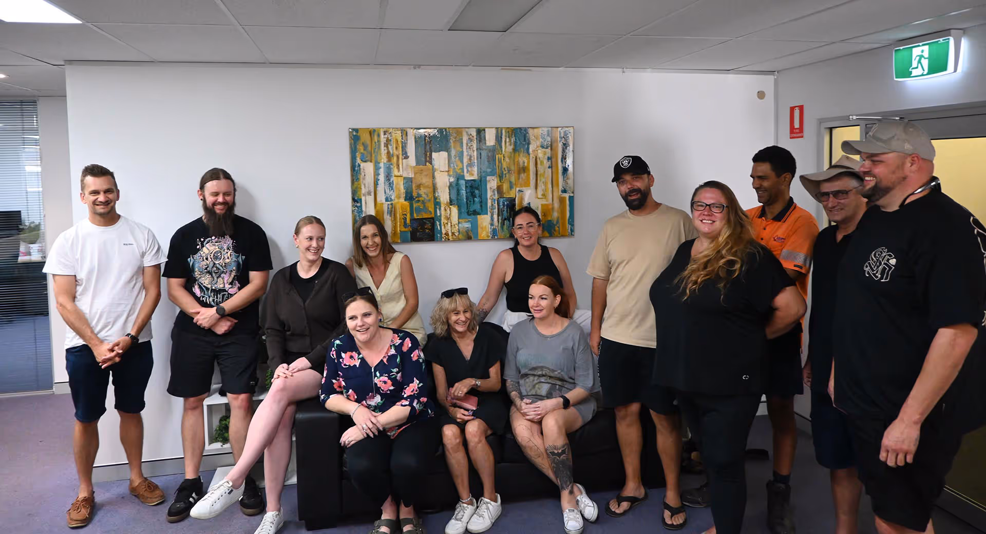 Group of eleven people smiling and posing in a modern office room with abstract wall art above a black couch.