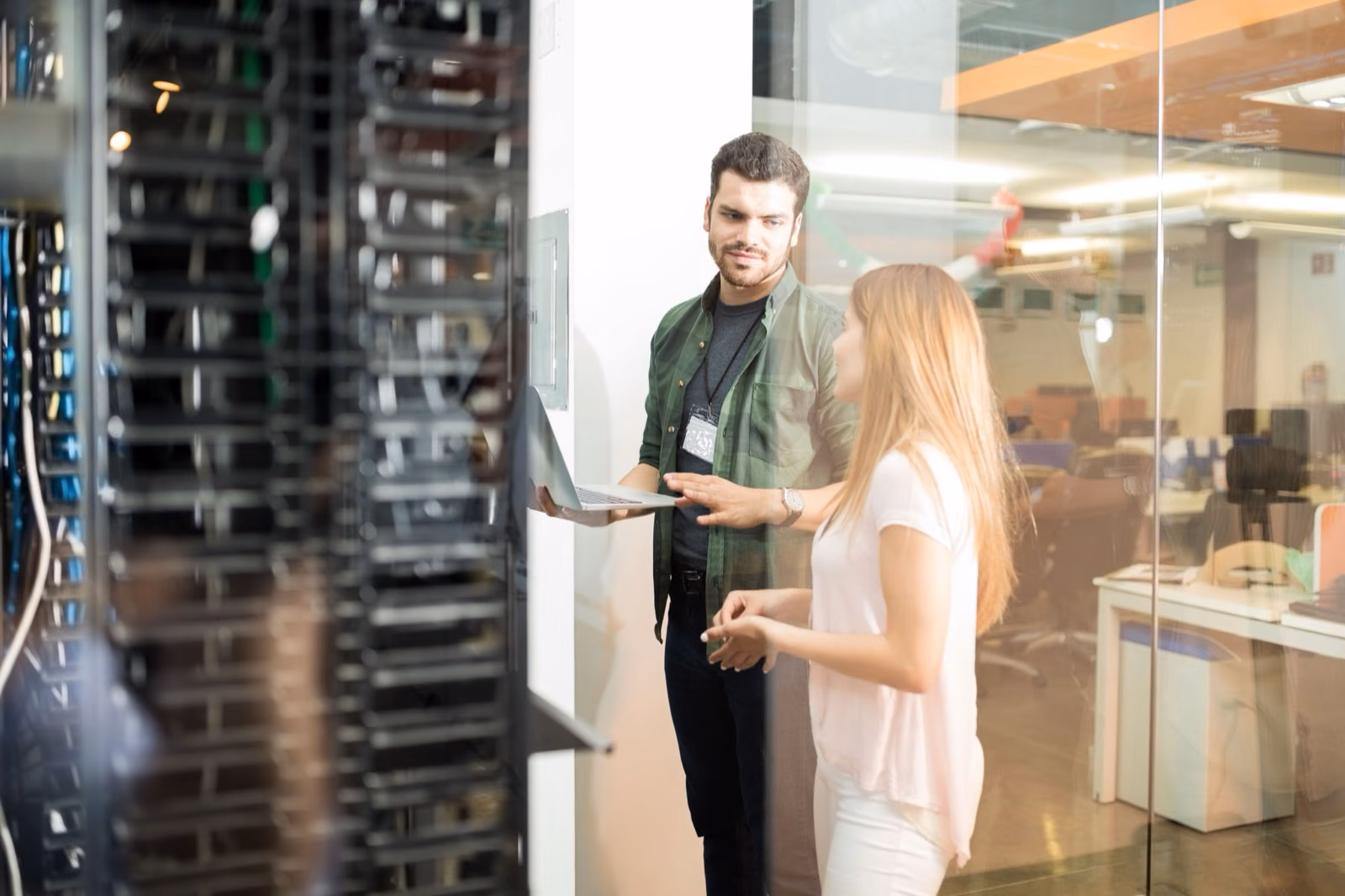 Two engineers talking with a laptop beside a glass-walled server room
