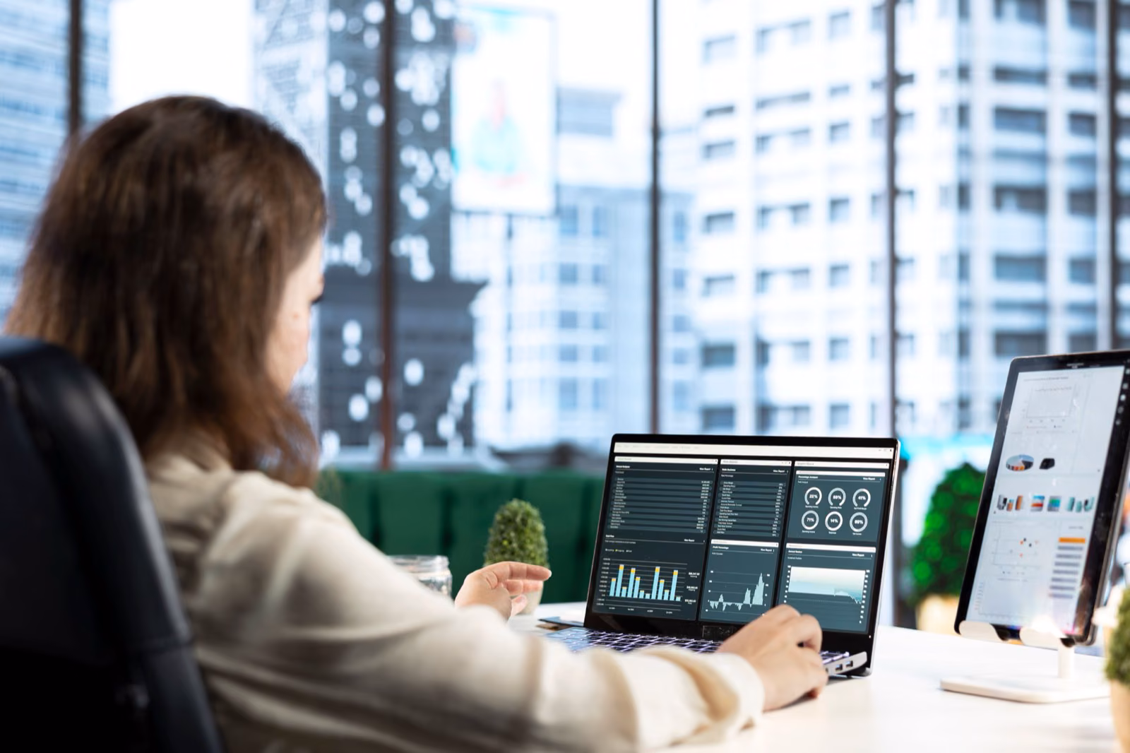 Finance analyst reconciling figures across a laptop and tablet dashboards by an office window