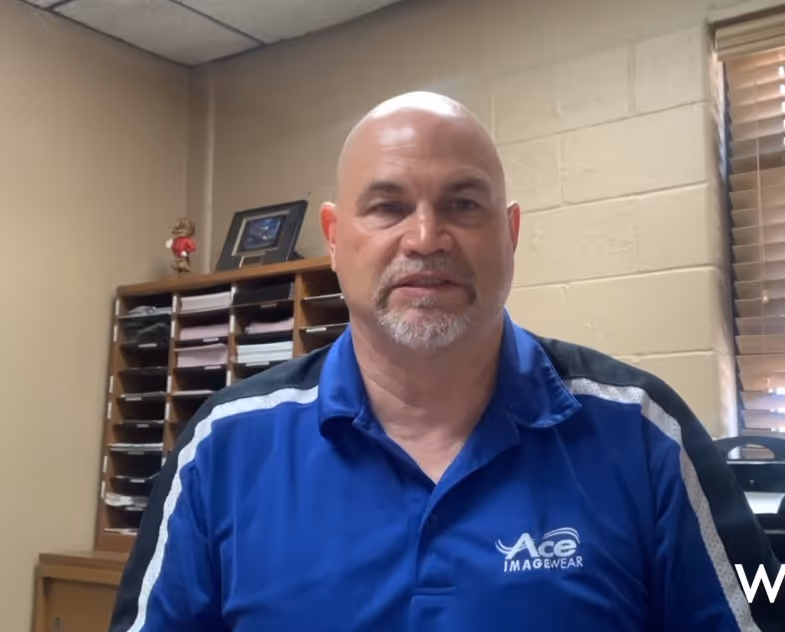 Bald man with a goatee wearing a blue Ace Imagewear polo shirt sitting in an office with mail slots and a window with blinds behind him.
