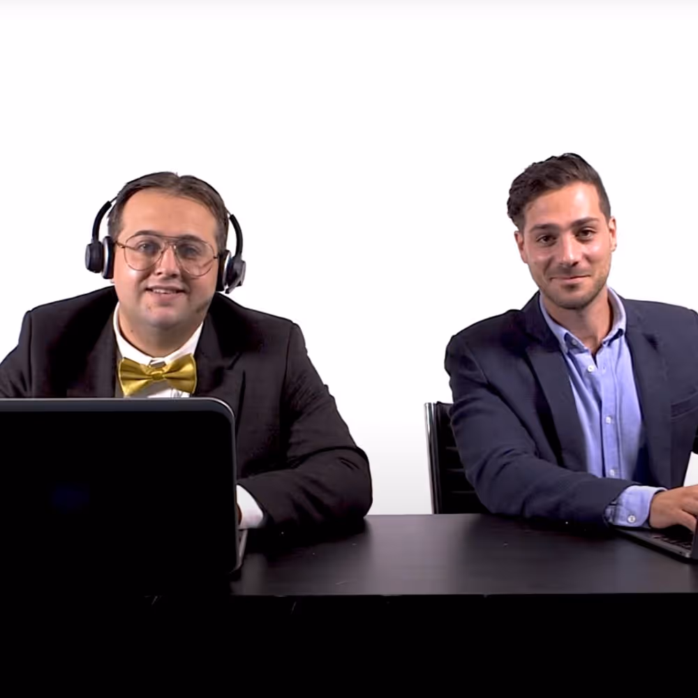 Two men sitting side by side at a black desk with laptops; one wearing a headset and yellow bow tie, the other in a dark blazer and blue shirt.