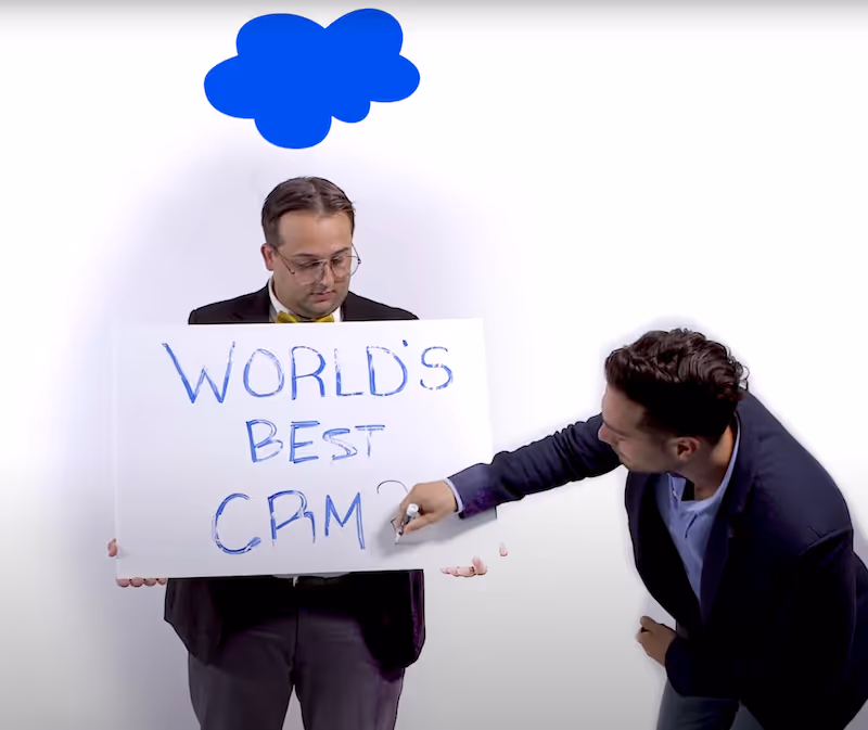 A man in glasses and a bow tie holds a sign reading 'WORLD'S BEST CRM?' while another man writes on the sign with a marker.