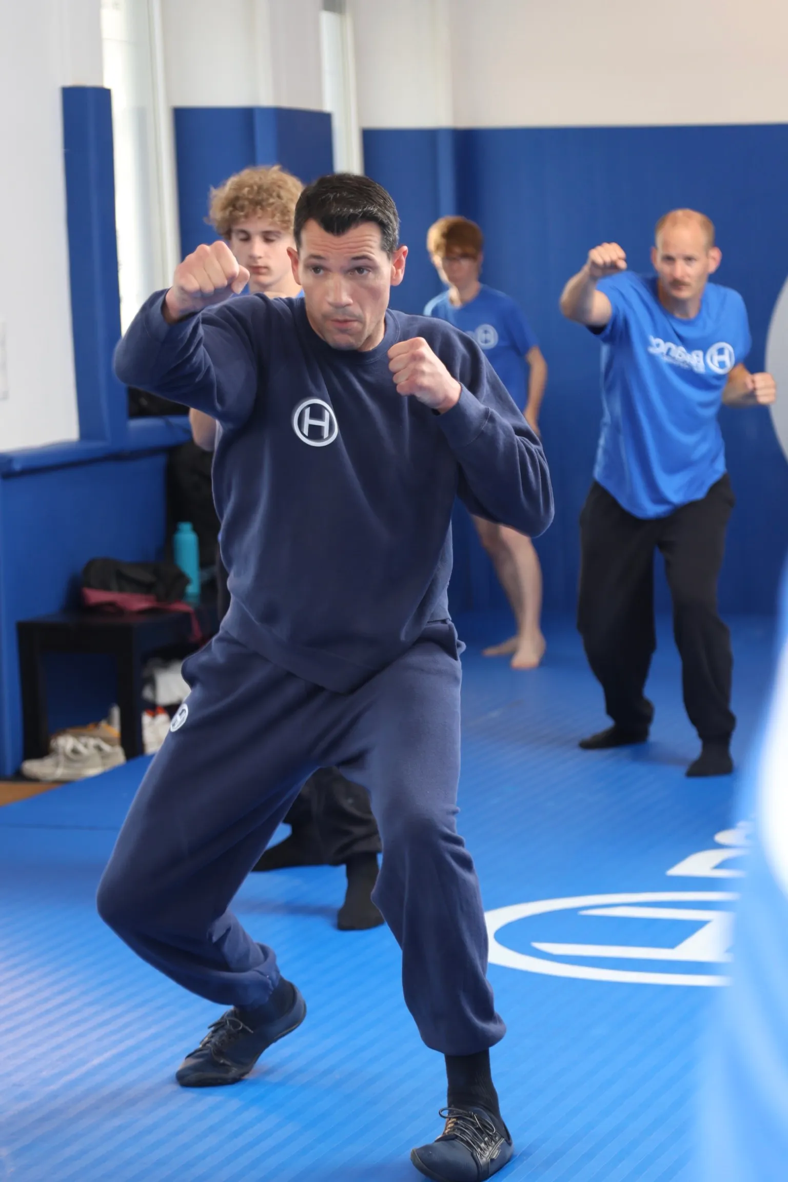 Instructeur H Balance en position de garde pendant un cours de self-défense à Strasbourg, avec des élèves qui s'entraînent en arrière-plan sur le tatami.