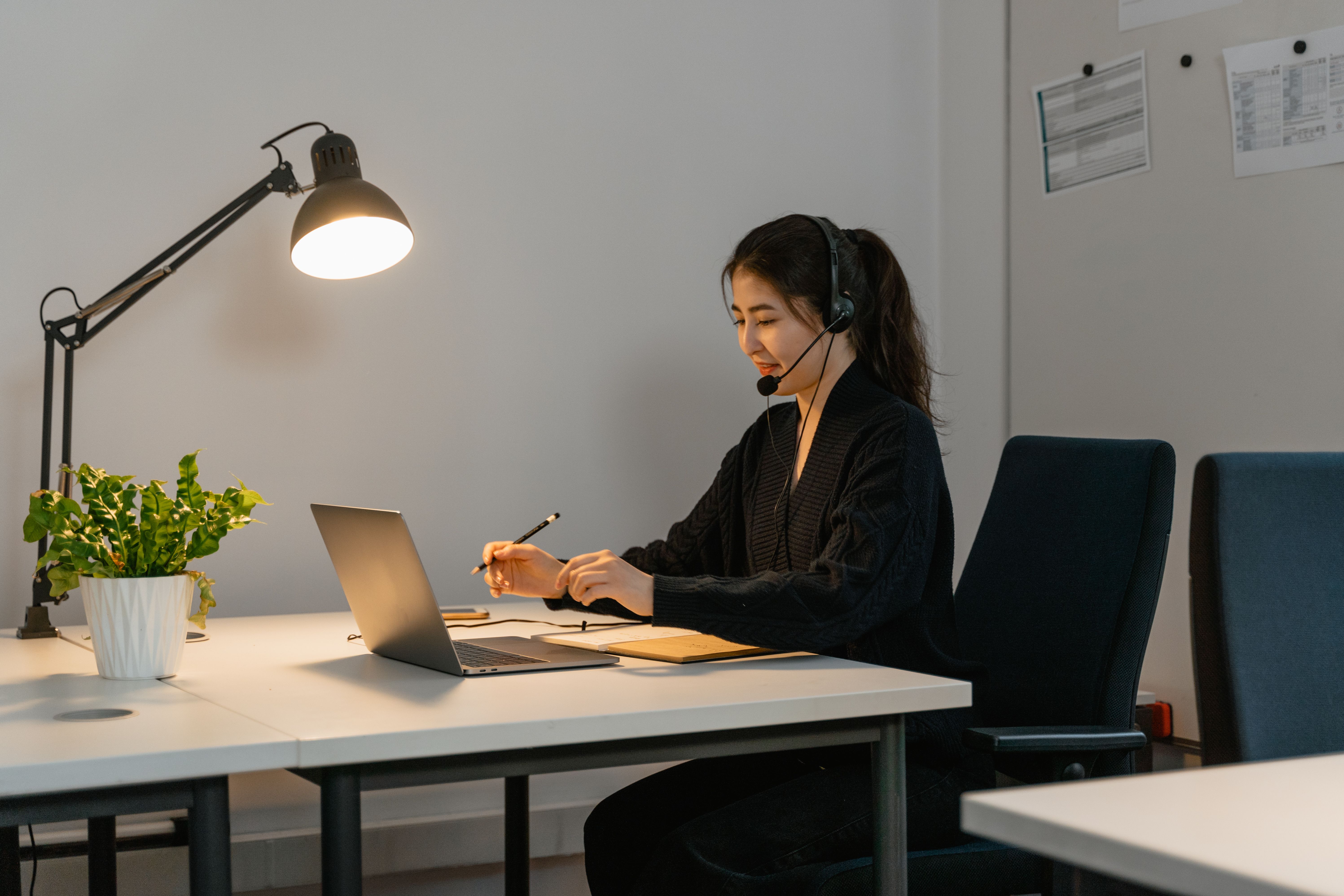Illustrative photograph of a headset-wearing support professional taking notes in an office, with her full face visible; photo by MART PRODUCTION on Pexels.