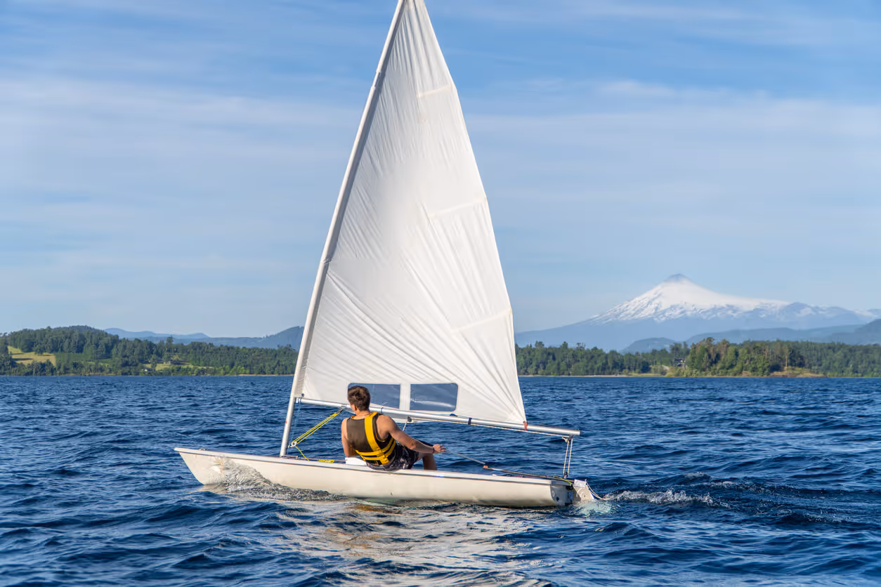 Man wearing a yellow life jacket sailing a small white dinghy with a large white sail on a lake with forested shoreline and a snow-capped mountain in the background.