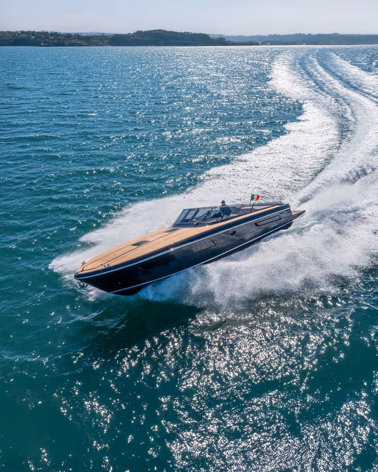 A sleek black and wood luxury speedboat cutting through sparkling ocean water near a coastline.