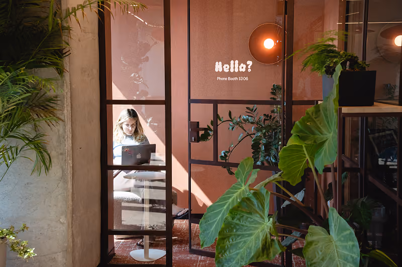 Woman working on a laptop inside a glass phone booth surrounded by plants.