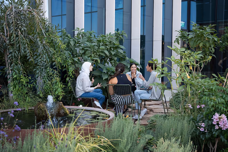 Four people sit and chat around a table near a small fountain in a lush garden courtyard.