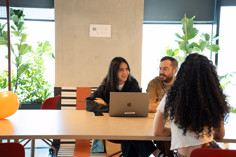 Three people having a discussion at a table with laptop and phone in a bright office with plants.