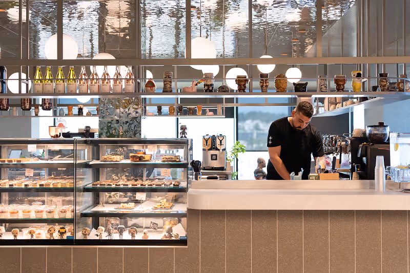 Barista preparing coffee behind a counter with pastries in glass displays and decorative mugs above.