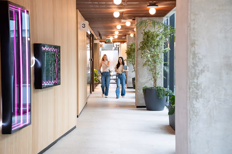 Two women walking and talking in a modern hallway with wooden walls and potted plants.
