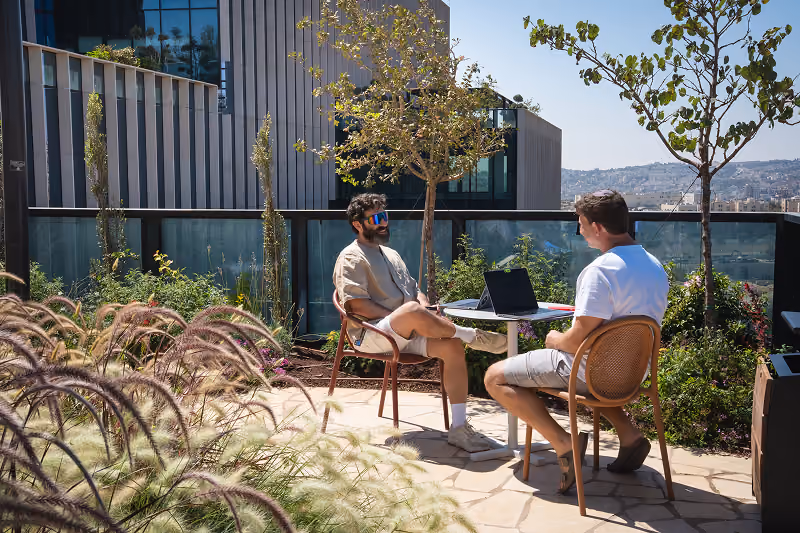 Two men sitting at a round table outdoors with a laptop, surrounded by greenery and cityscape.