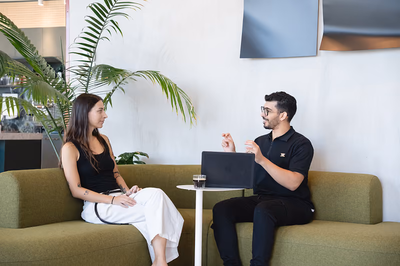 Man and woman sitting on green couches, talking across a small table with a laptop and a glass of drink.