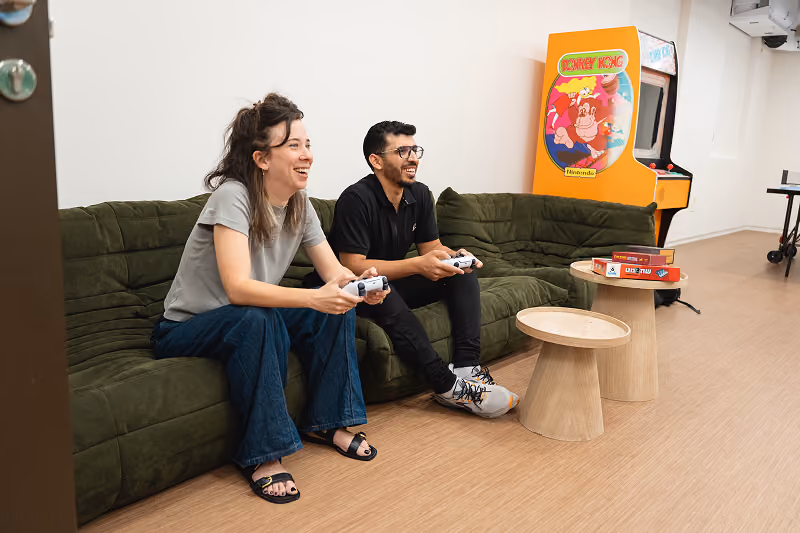 Two people sitting on a green couch playing video games with controllers in a room with Donkey Kong arcade.