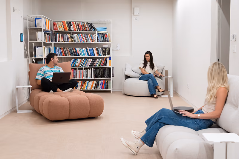 Three people sitting in a modern room with laptops, bookshelves, and soft seating furniture.