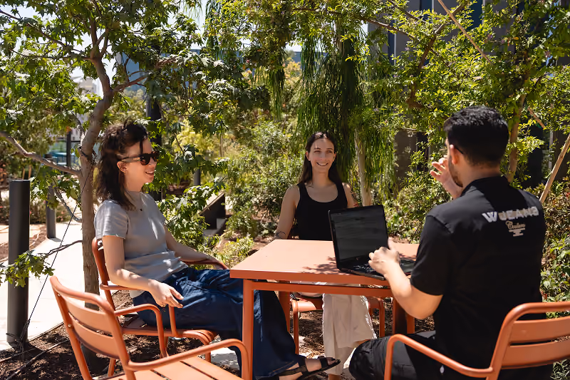 Three people sitting at an outdoor table having a conversation with laptop in a sunny garden setting.