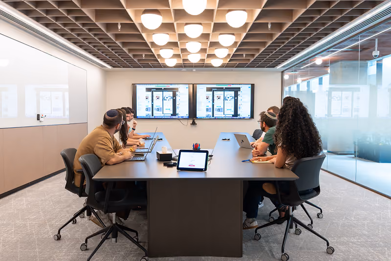 Five people working on laptops in a modern conference room, looking at two digital screens on the wall.