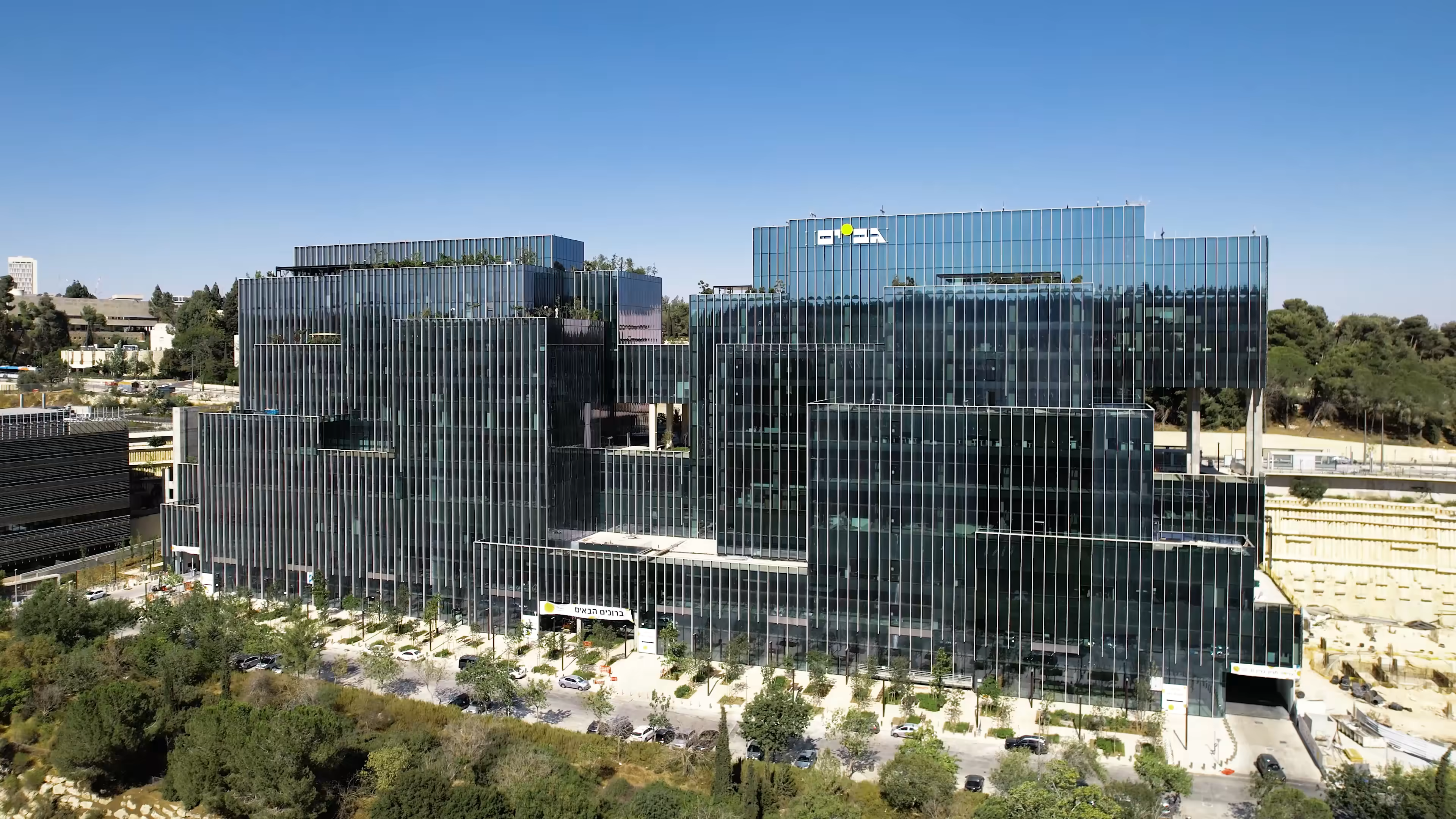 Modern glass office complex with multiple connected buildings surrounded by trees and a parking area.