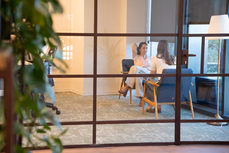 Two women having a conversation while sitting on blue chairs in a modern office room.