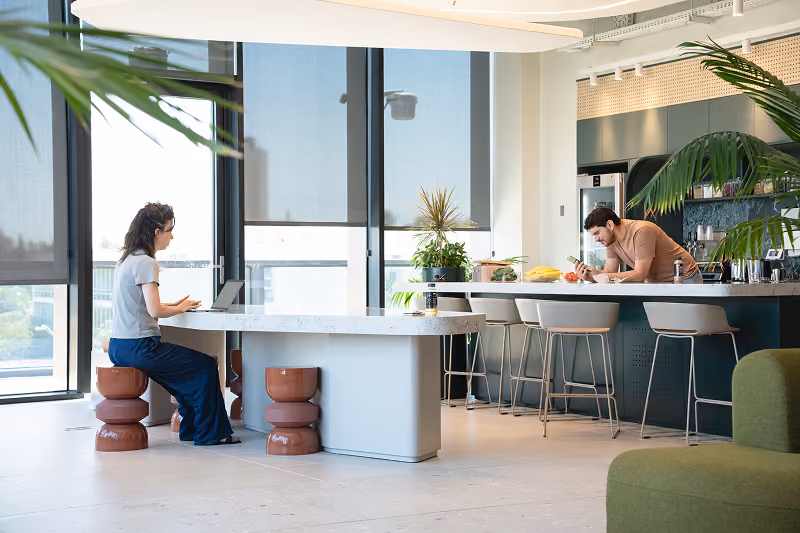Two people working in bright modern kitchen with large windows, plants, and bar seating.