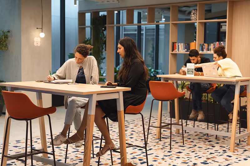 Four people sitting at two tables in a modern, bright workspace with books and plants.