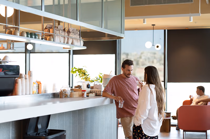 Two people chatting and holding drinks near a cafe counter with a seated man in the background.