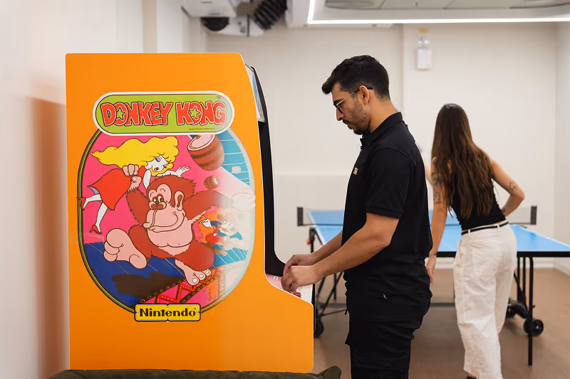 Man playing on a vintage Donkey Kong arcade machine while a woman stands near a ping pong table.