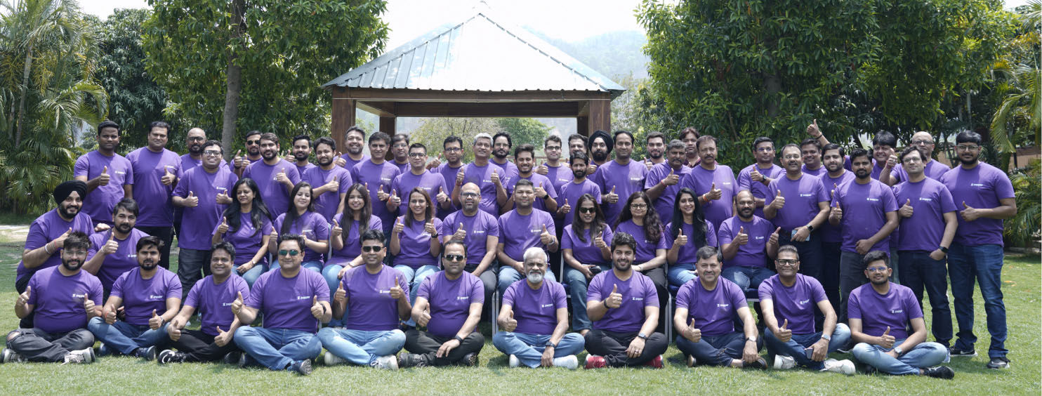 Large group of people wearing matching purple t-shirts posing outdoors on grass in front of a wooden gazebo.
