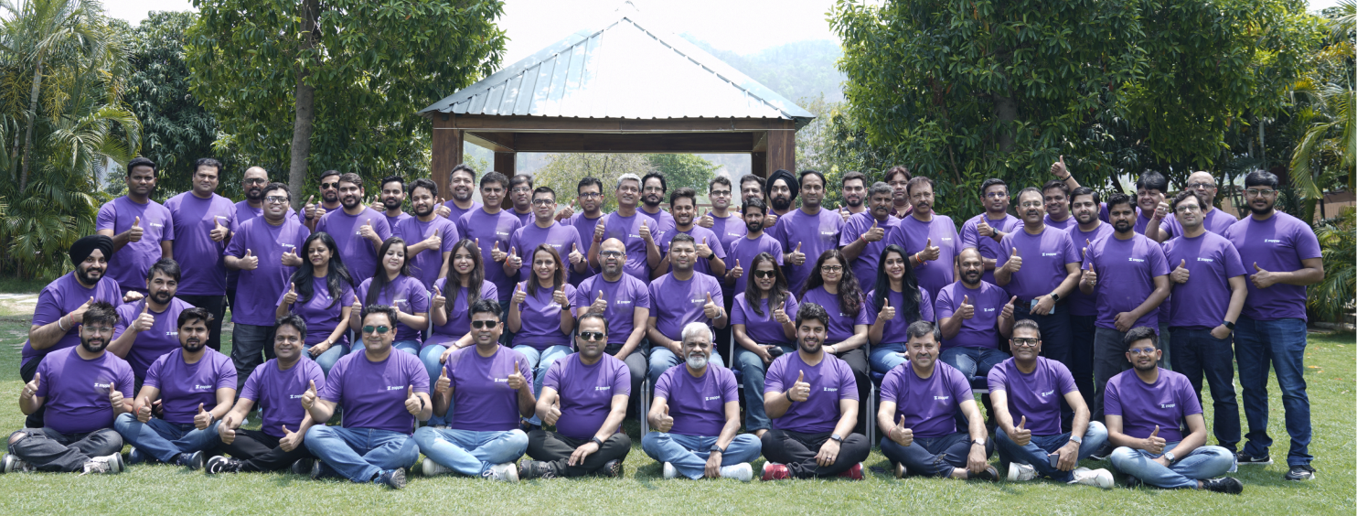 Large group of people wearing matching purple t-shirts posing outdoors on grass in front of a wooden gazebo.