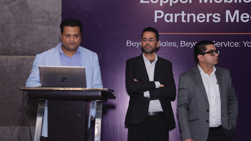 Three men in business attire standing on a stage during a presentation or meeting; one man is behind a podium with a laptop, the other two are standing with arms crossed or hands in pockets.