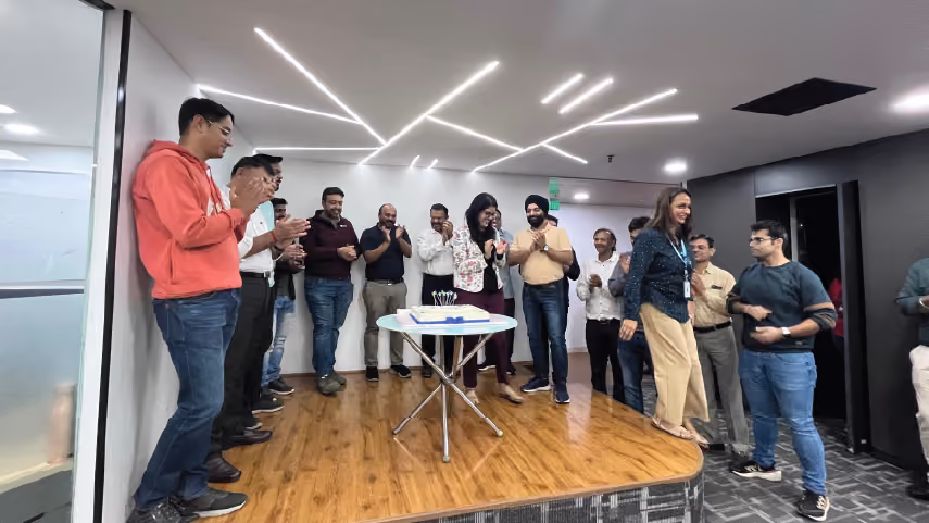 Group of colleagues standing on a small stage in an office, clapping and celebrating around a table with a cake.
