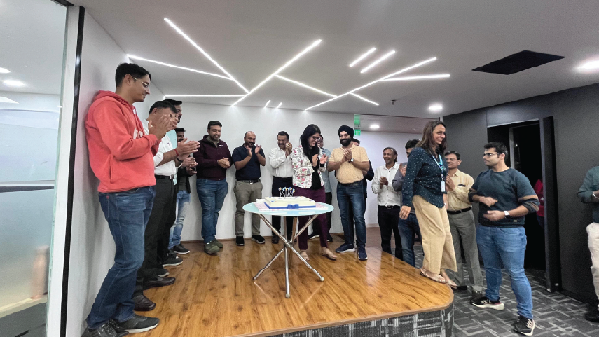 Group of colleagues standing on a small stage in an office, clapping and celebrating around a table with a cake.