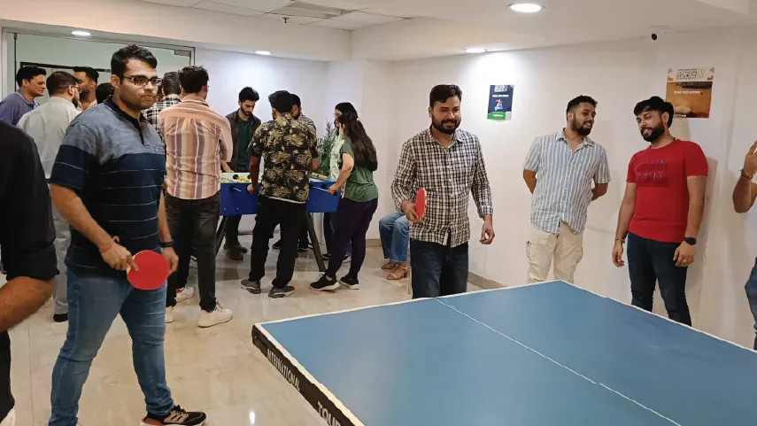 Group of people playing table tennis and foosball in a room with white walls and tiled floor.