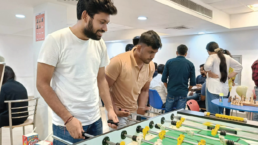 Two men playing foosball in a room with several people in the background engaged in conversation.