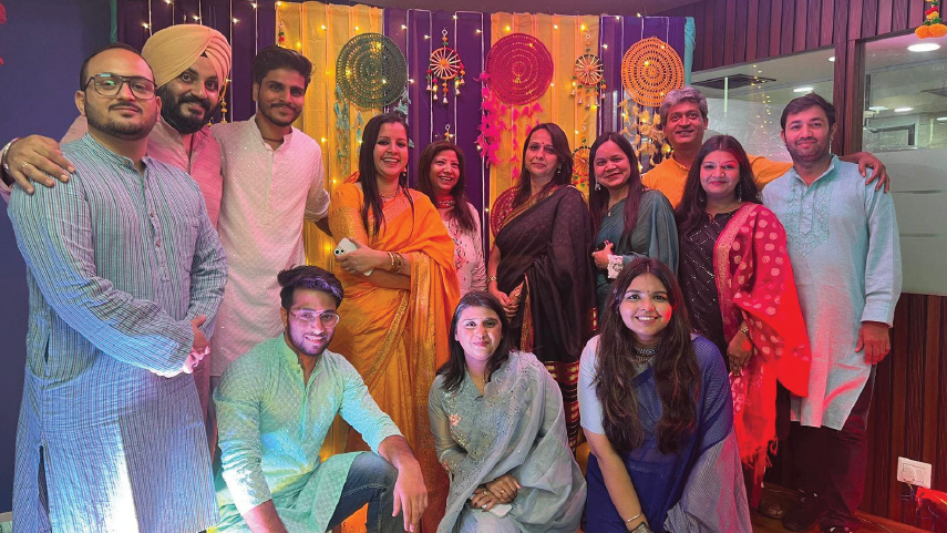 Group of thirteen people in traditional Indian attire posing indoors with colorful decorative hangings and lights in the background.