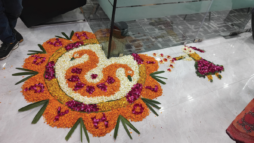 Colorful flower rangoli design with orange, white, and purple petals, featuring a central artistic pattern and green leaves, placed on a tiled floor near glass walls.