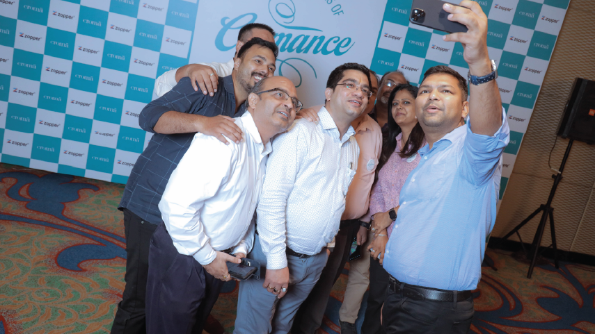 Group of six adults smiling and posing for a selfie at an indoor event with branded banners in the background.