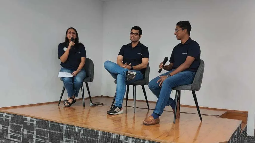 Three people sitting on chairs on a stage, two men and one woman, holding microphones during a discussion or panel event.