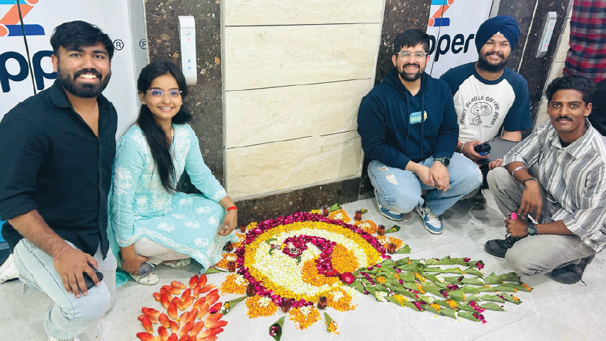 Five people sitting on the floor smiling around a colorful flower rangoli with leaf and flower decorations, indoors near a wall with a logo.