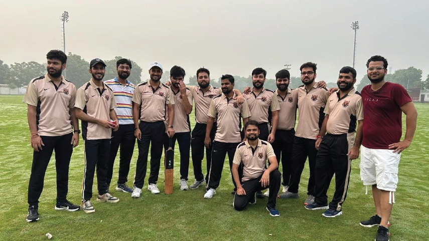 Group of twelve men posing on a grassy field, most wearing matching beige cricket uniforms with black pants, one holding a cricket bat.