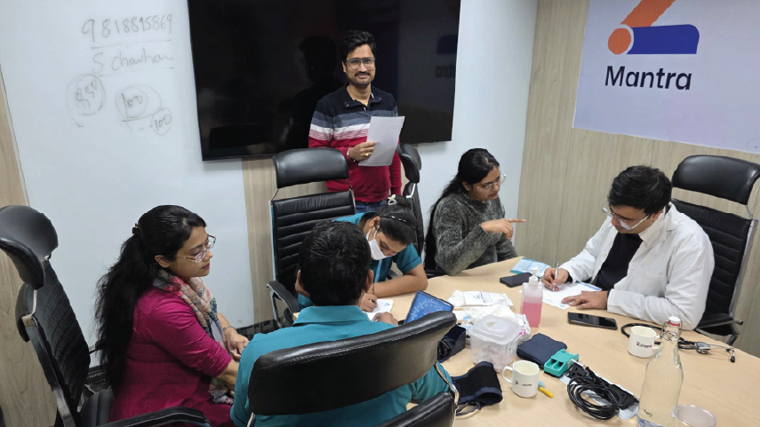 A group of six people in an office meeting room with a whiteboard and a Mantra logo on the wall, some are seated and writing while one person stands holding a paper.