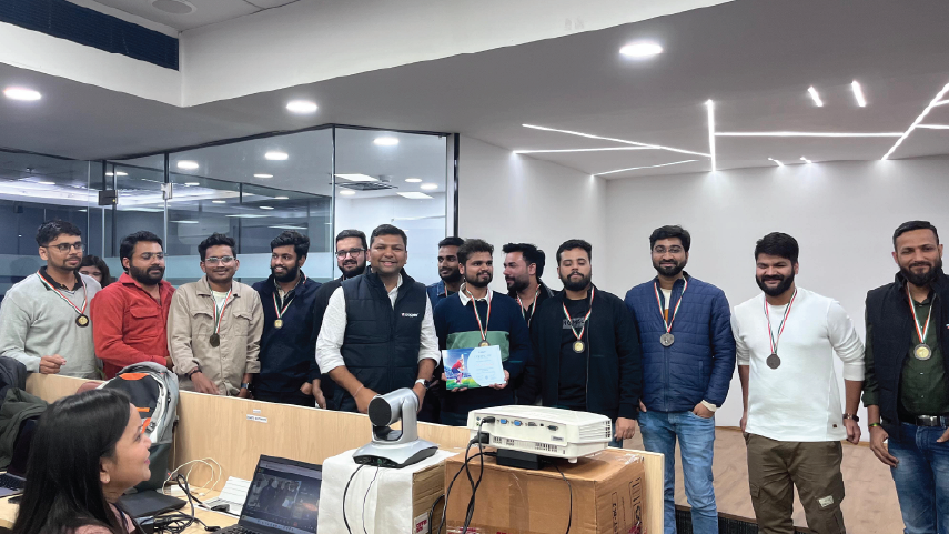 Group of men standing in an office wearing medals, with one man holding a certificate, smiling near a desk with a laptop and projector.