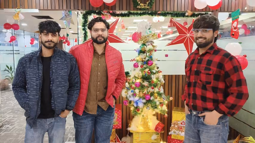 Three men standing indoors around a decorated Christmas tree with festive red and white balloons and holiday decorations in the background.