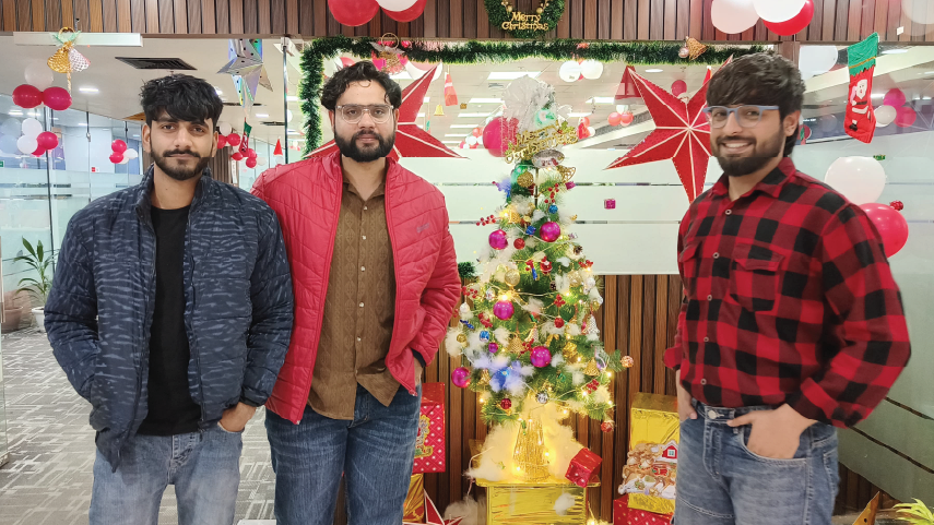 Three men standing indoors around a decorated Christmas tree with festive red and white balloons and holiday decorations in the background.