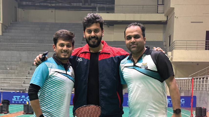 Three men posing and smiling together on an indoor badminton court, with two holding badminton rackets.
