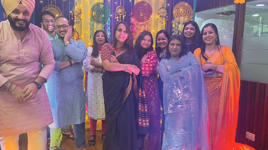 Group of nine people wearing colorful traditional Indian attire posing indoors with decorative dreamcatchers hanging in the background.