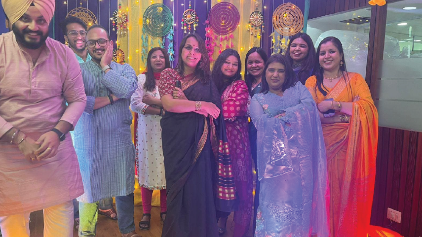 Group of nine people wearing colorful traditional Indian attire posing indoors with decorative dreamcatchers hanging in the background.