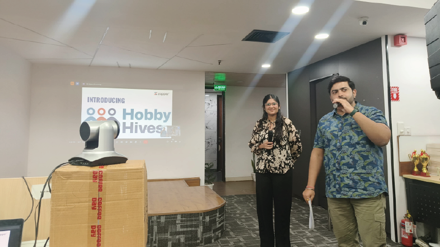 Two people standing and holding microphones in an office room, with a projector displaying 'Introducing Hobby Hives' on the wall behind them.