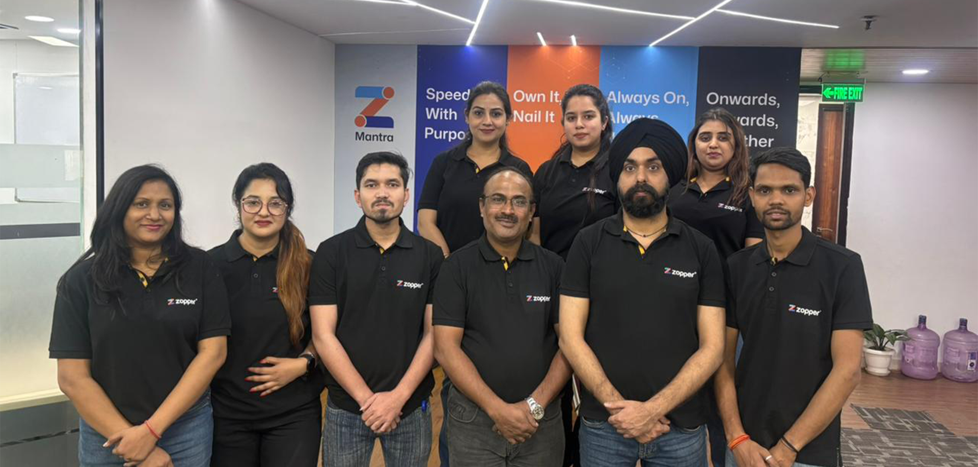 Group photo of nine men and women wearing black Zopper polo shirts standing in an office with a colorful Mantra poster behind them.