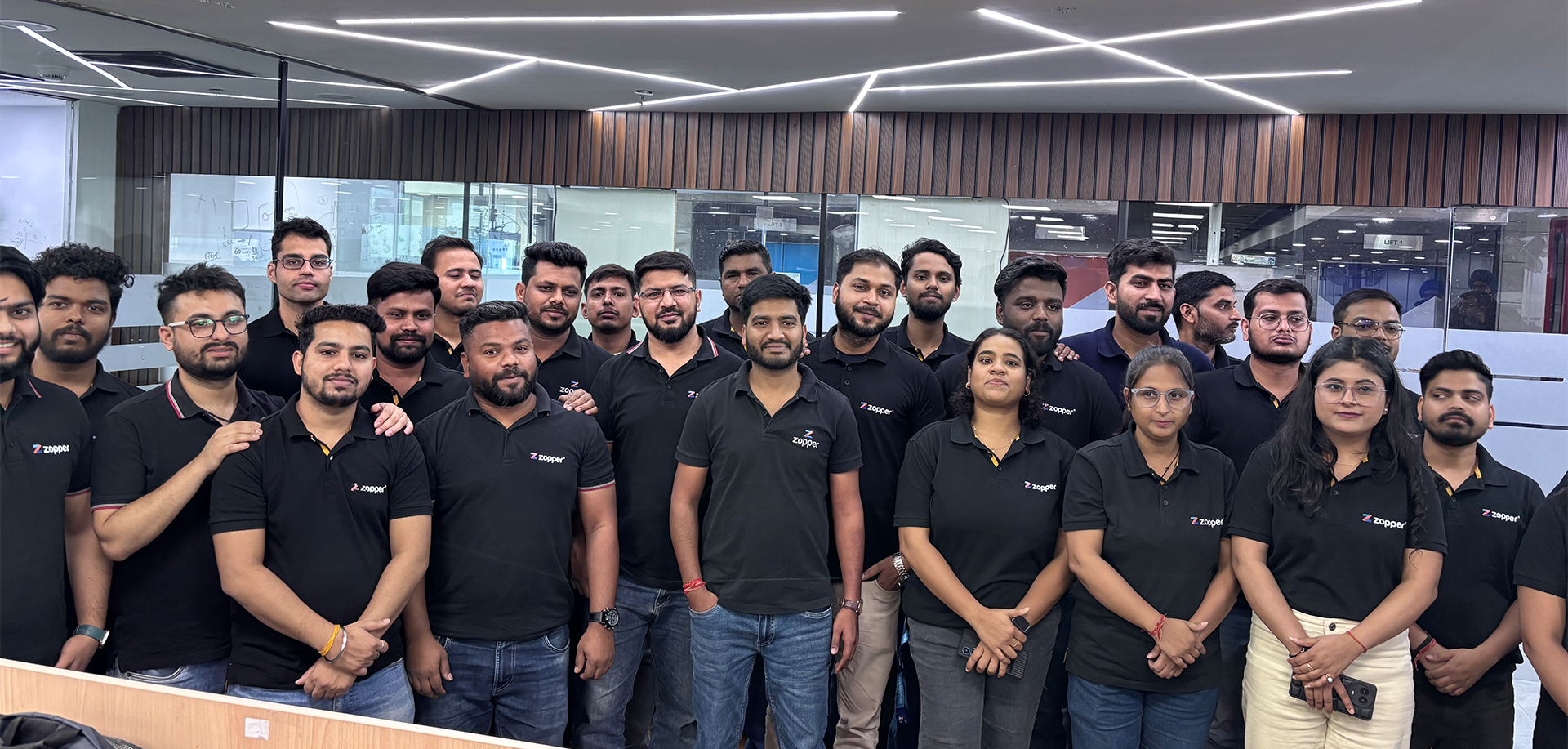 Group of men and women wearing black Zopper polo shirts posing inside a modern office with a wooden panel wall and glass partitions.